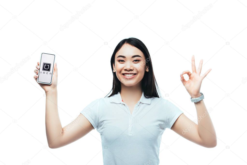 KYIV, UKRAINE - JULY 15, 2019: happy brunette asian woman holding smartphone with uber app and showing ok sign isolated on white