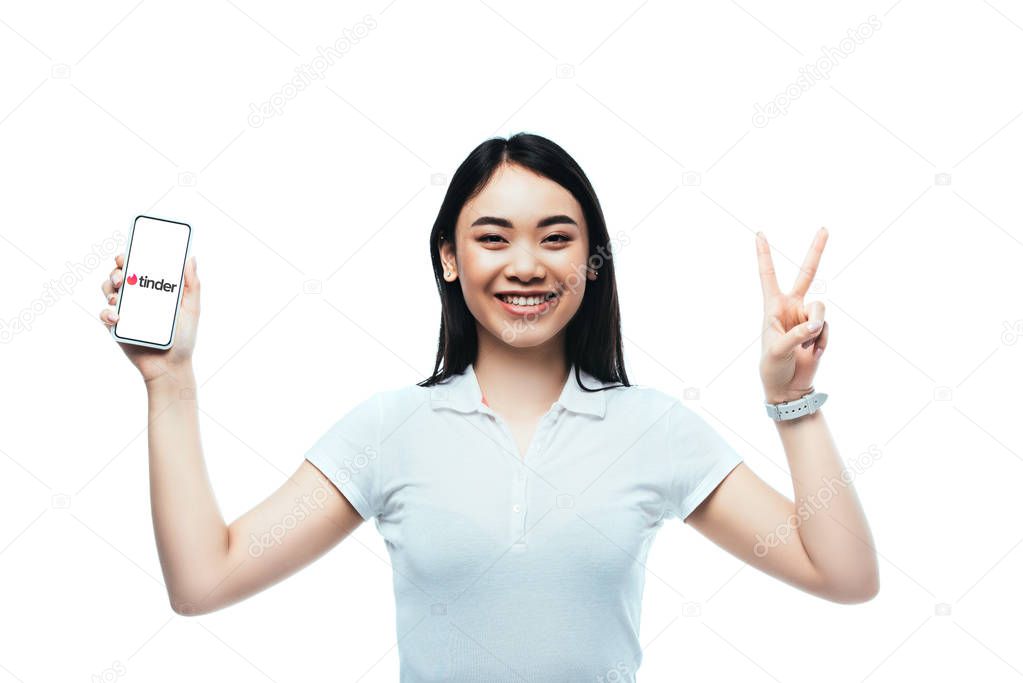 KYIV, UKRAINE - JULY 15, 2019: happy brunette asian woman holding smartphone with tinder app and showing peace sign isolated on white