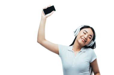Gülümseyen, kulaklıklı çekici Asyalı kız elinde beyaz ekranlı akıllı bir telefon tutuyor.