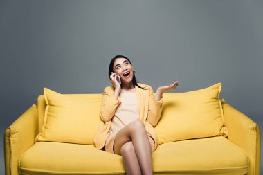 happy asian businesswoman sitting on yellow sofa with crossed legs and talking on smartphone on grey background clipart