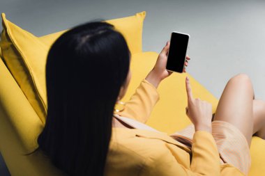 Esmer iş kadını sarı koltukta oturuyor ve parmağıyla gri ekranlı akıllı telefonu işaret ediyor.