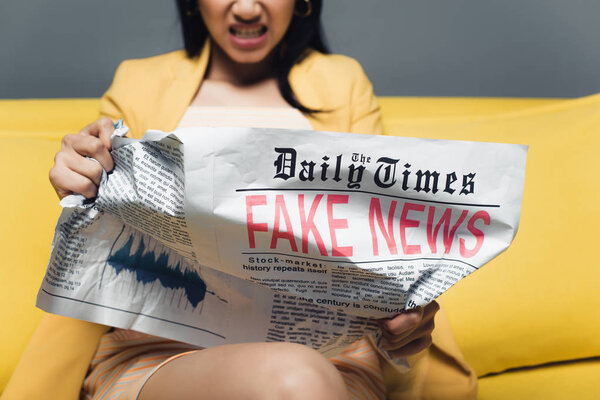 cropped view of angry asian businesswoman sitting on yellow sofa and reading newspaper with fake news on grey background