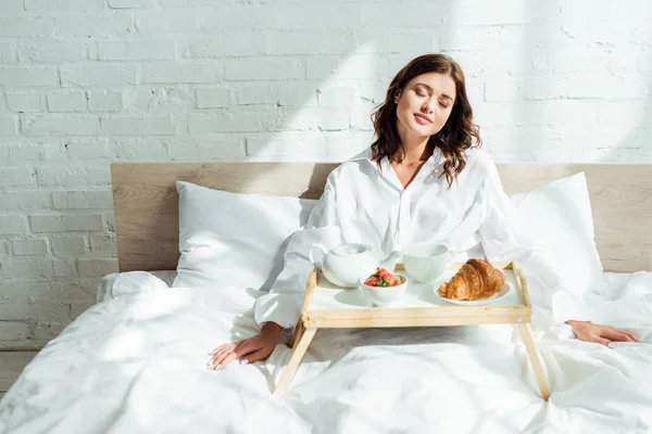 早上穿着白衬衫躺在床上吃早餐的漂亮女人 — 图库照片