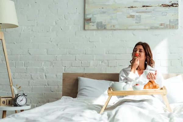 早上穿着白衬衫躺在床上吃早餐的漂亮女人 — 图库照片