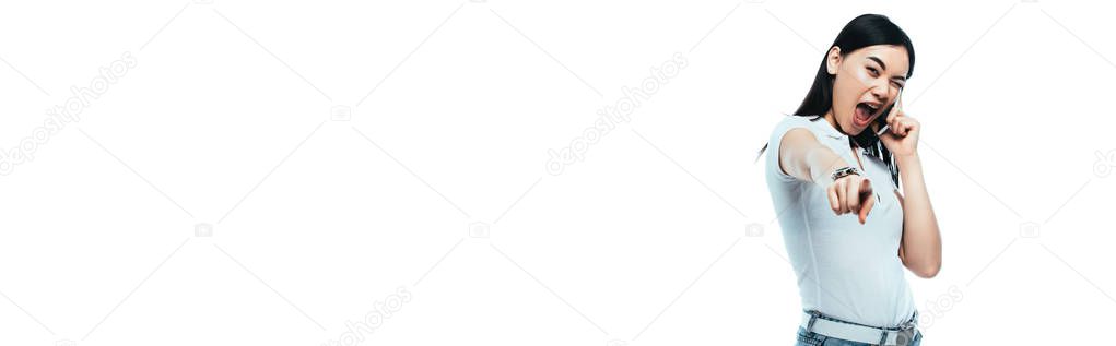 excited brunette asian woman talking on smartphone and pointing with finger at camera isolated on white, panoramic shot
