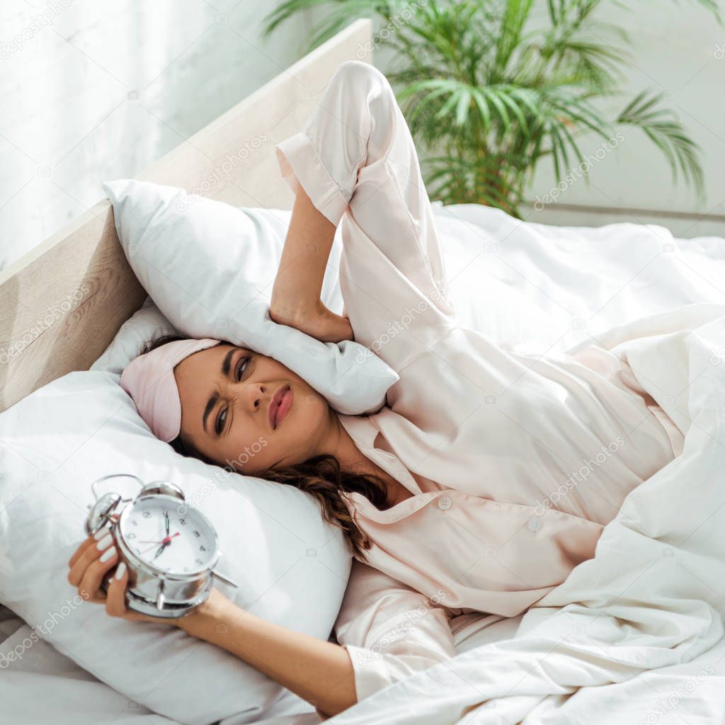 angry woman in sleeping mask holding alarm clock at morning 
