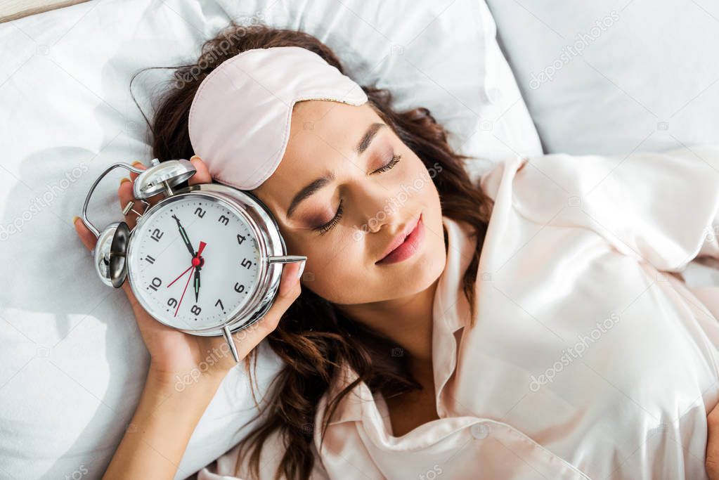 top view of attractive woman with sleeping mask holding alarm clock at morning 