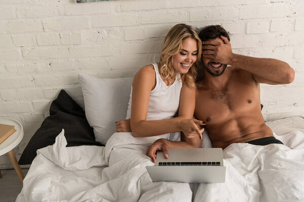 laughing couple pointing at laptop in bed in the morning