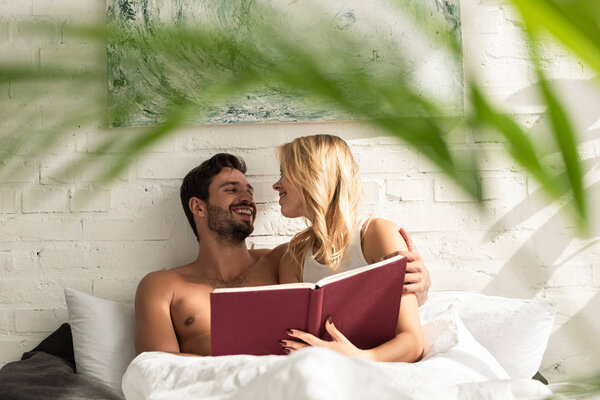 happy couple hugging and reading book together in bed in the morning, selective focus of plant