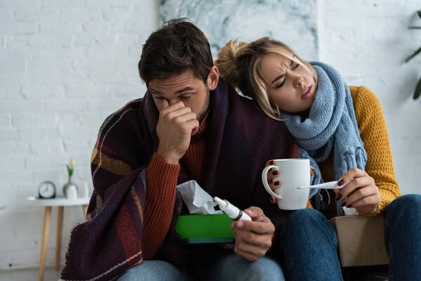 diseased couple holding thermometer, hot drink and nasal spray in bedroom