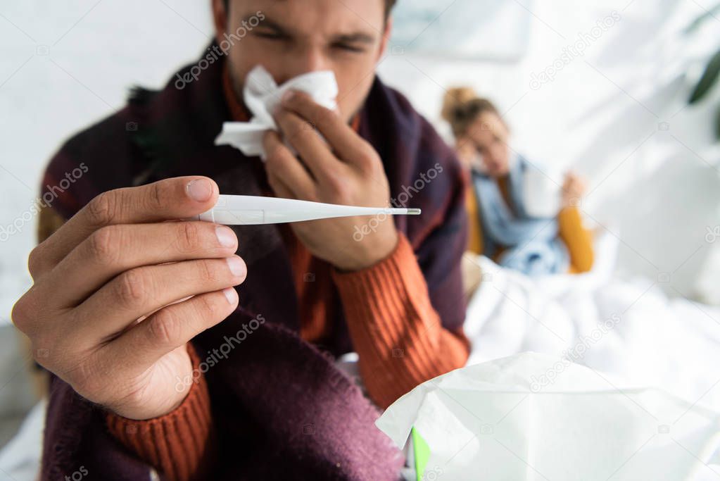 selective focus of sick man with fever holding thermometer and napkin in bed with woman behind