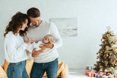 Mutlu ebeveynler süslenmiş Noel ağacının yanında duruyor ve ellerinde sevimli bir bebek var.