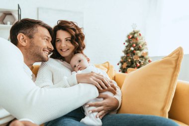 Mutlu adam karısına sarılıyor, kanepeye oturuyor ve kucağında sevimli bir bebek var.