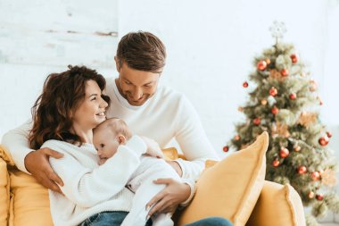 Mutlu adam, karısına sarılıyor. Sarı koltukta oturuyor. Yanında bebek, Noel ağacının yanında.
