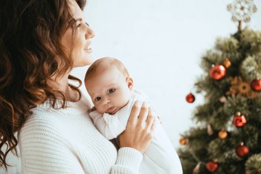 Mutlu anne, Noel ağacının yanında dururken tapılası bebeği tutuyor.