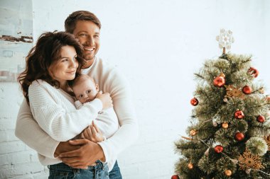 Mutlu adam, Noel ağacının yanında dururken karısını bebeğiyle kucaklıyor.