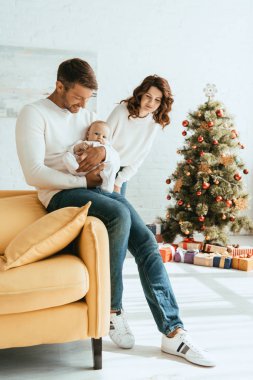 Mutlu kadın Noel ağacının yanında duruyor ve kucağında tatlı bir bebek olan kocaya bakıyor.
