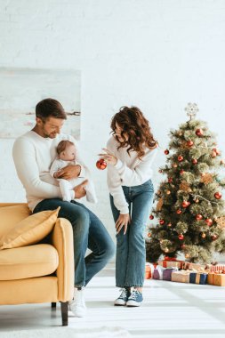 Anne bebeğe noel balosunu gösteriyor. Babaların elleri süslemeli Noel ağacının yanında oturuyor.