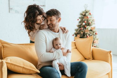 Mutlu bir kadın, sarı koltukta oturan kocasının omzunun üzerinden sevimli bir bebeğe bakıyor.