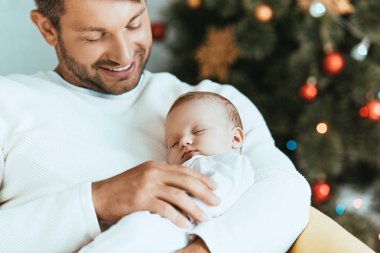 Gülümseyen baba Noel ağacının yanında uyuyan bebeği tutuyor.