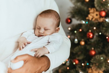 Babamın uyuyan bebeği Noel ağacının yanında tutarken çekilmiş görüntüsü.