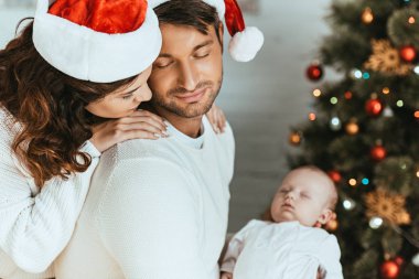 Mutlu kadın kocasının omzunun üzerinden uyuyan bebeğe bakıyor.