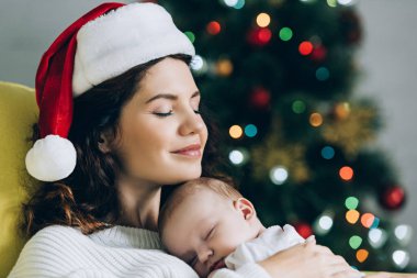 Noel Baba şapkalı mutlu kadın gözleri kapalı ve kucağında uyuyan bir bebek.