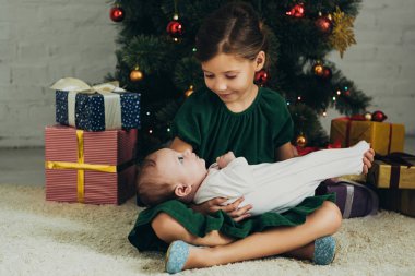 Gülümseyen çocuk Noel ağacının ve hediye kutularının yanında otururken kucağında şirin bir bebek tutuyor.