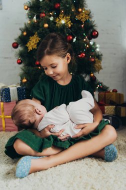 Noel ağacının ve hediye kutularının yanında otururken kucağında küçük şirin bir kız çocuğu.