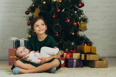 Mutlu çocuk Noel ağacının ve hediye kutularının yanında otururken kucağında şirin bir bebek tutuyor.