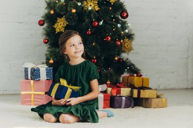 Mutlu çocuk Noel ağacının yanında oturuyor, hediye kutusunu tutuyor ve başka tarafa bakıyor.