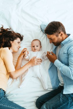 happy parents gently touching adorable baby while lying on white bedding together clipart