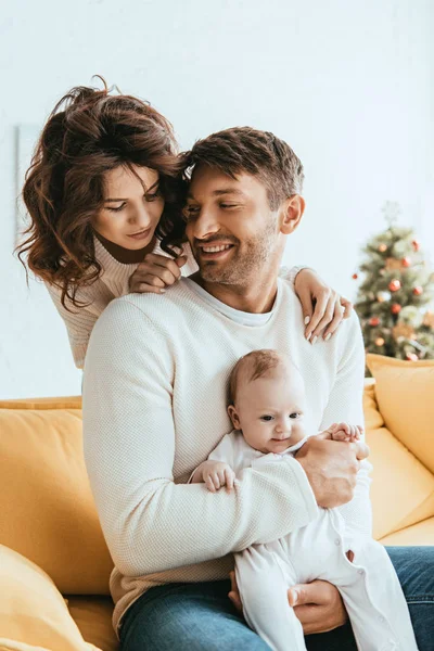 Güzel bir kadın, sarı koltukta oturan kocasının omzunun üzerinden şirin bir bebeğe bakıyor.