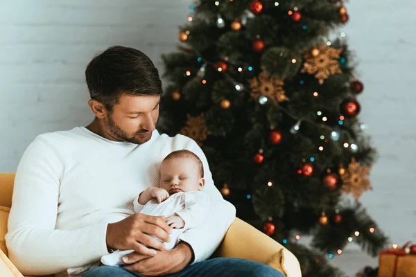 幸せな父親はクリスマスツリーの近くに座っている間に眠っている赤ちゃんを保持 — ストック写真