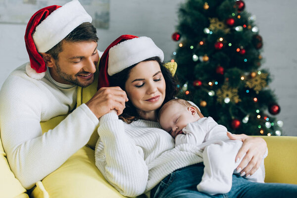 счастливый мужчина трогает руку улыбающейся жены, держащей спящего ребенка
