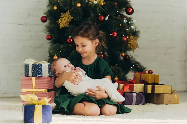 Criança Adorável Segurando Bebê Bonito Enquanto Sentado Perto Árvore Natal — Fotografia de Stock