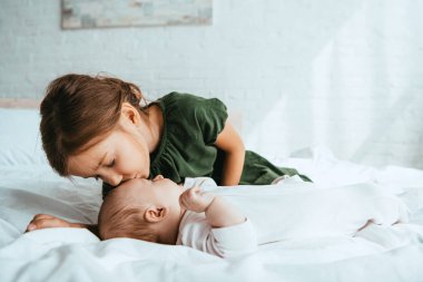 Küçük kız kardeşini öpen sevimli çocuk beyaz yatağa uzanıyor.