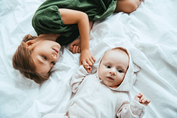 top view of smiling child holding hand of little sister lying on white bedding