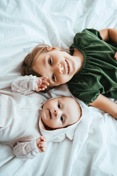 top view of smiling child looking at camera while lying on white bedding near little sister