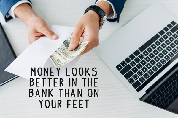 top view of businessman holding envelope with dollar banknotes near laptop on table with money looks better in the bank than on your feet illustration