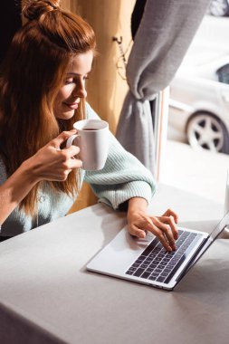 beautiful redhead woman with cup of coffee using laptop in cafe