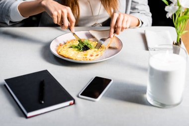 Kahvede kahvaltıda omlet yiyen, not defteri ve akıllı telefonlu bir kadın manzarası.