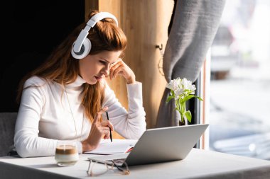 Kulaklıklı güzel, düşünceli bir kadın kafede dizüstü bilgisayarda Webinar izliyor.