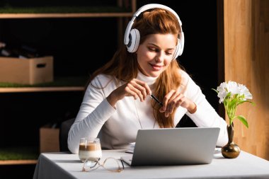 kızıl saçlı, kulaklıklı bir kadın kafede dizüstü bilgisayarda Webinar izliyor.