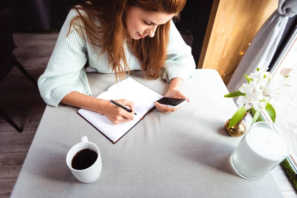 Mulher Atraente Usando Smartphone Escrevendo Bloco Notas Café Com Xícara — Fotografia de Stock