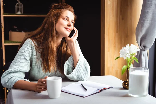 笑顔の女性がノートとコーヒーを片手にカフェでスマホで話しています — ストック写真