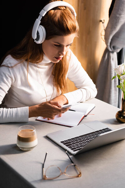 концентрированная женщина в наушниках, записывающая в блокнот во время просмотра вебинара на ноутбуке в кафе со стаканом кофе
