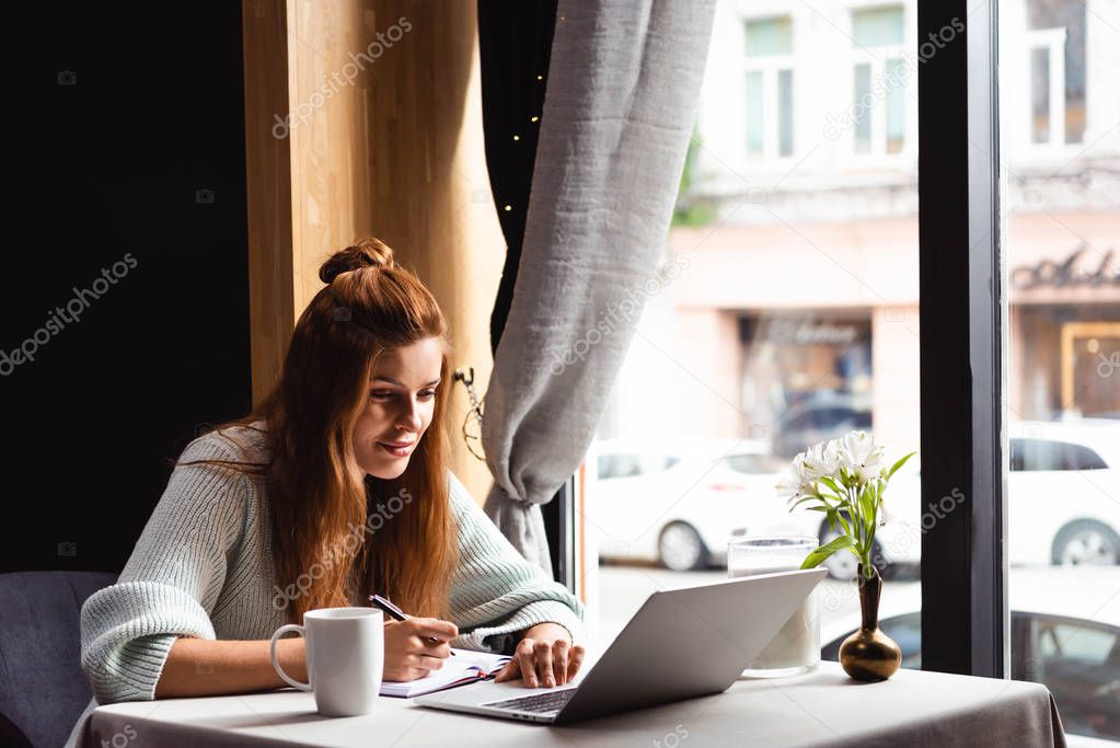 smiling woman writing in notepad while watching webinar on laptop in cafe