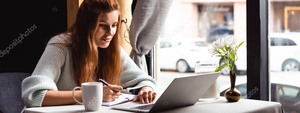 attractive woman writing in notepad while watching webinar on laptop in cafe