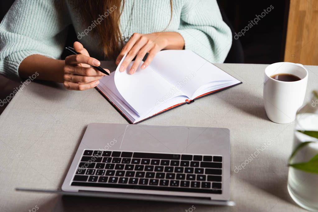 cropped view of woman studying online with notepad and laptop in cafe with cup of coffee  